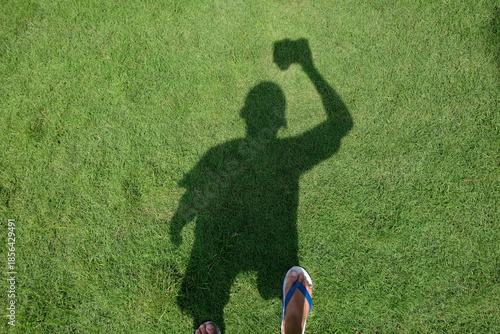 Shadow of a person holding a camera cast on green grass lawn, creating a creative self-portrait concept with natural light, minimal composition, and outdoor photography mood.