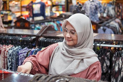 Asian hijab woman browsing beautiful traditional Indonesian batik clothes in a vibrant local boutique. Authentic cultural fashion, ethnic patterns, and retail shopping lifestyle