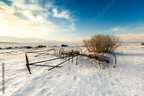 Vintage hay rake abandoned in snow at Lake Cildir, Kars, Turkey. Rustic agricultural machinery on frozen shore during winter golden hour with mountains.