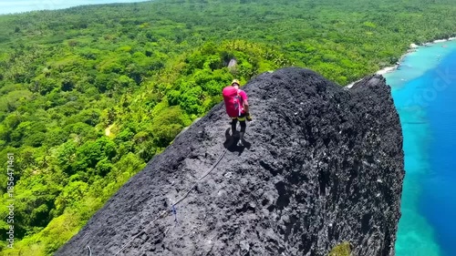 A climber on a dark rock ridge overlooks dense green forests and blue sea