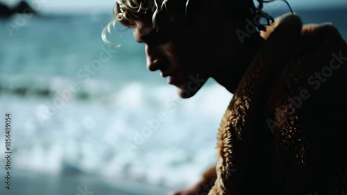 A man stands by the ocean during sunset, gazing thoughtfully towards the water. The waves crash gently against the shore, creating a tranquil atmosphere. His expression shows deep reflection and calm