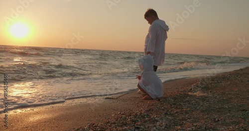 A brother and sister enjoy an evening at the beach, drawing in the wet sand with a stick. The children run away from a small wave. The children watch a beautiful golden sunset over the calm sea.