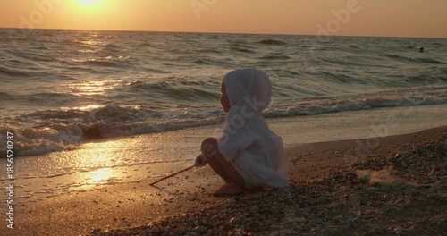 The child plays on the seashore during sunset. A small child plays with a stick on a wet sand, when delicate ocean waves I run ashore during a beautiful gold sunset, enjoying calm summer holidays.