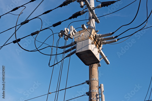 青空にそびえる電柱最上部の高圧柱上開閉器と配線、電力供給インフラの風景