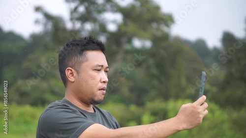 Man holding a smartphone while recording or taking photos in an open area with trees in the background.
