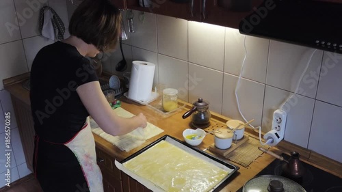 Woman meticulously preparing phyllo pastry dough in a cozy home kitchen. She's layering sheets on a wooden counter, ready for baking, capturing the essence of homemade culinary creation.