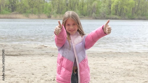Smiling girl in a pink jacket stands on a sandy riverbank, forming a like shape with her hands against a calm water and green forest backdrop.