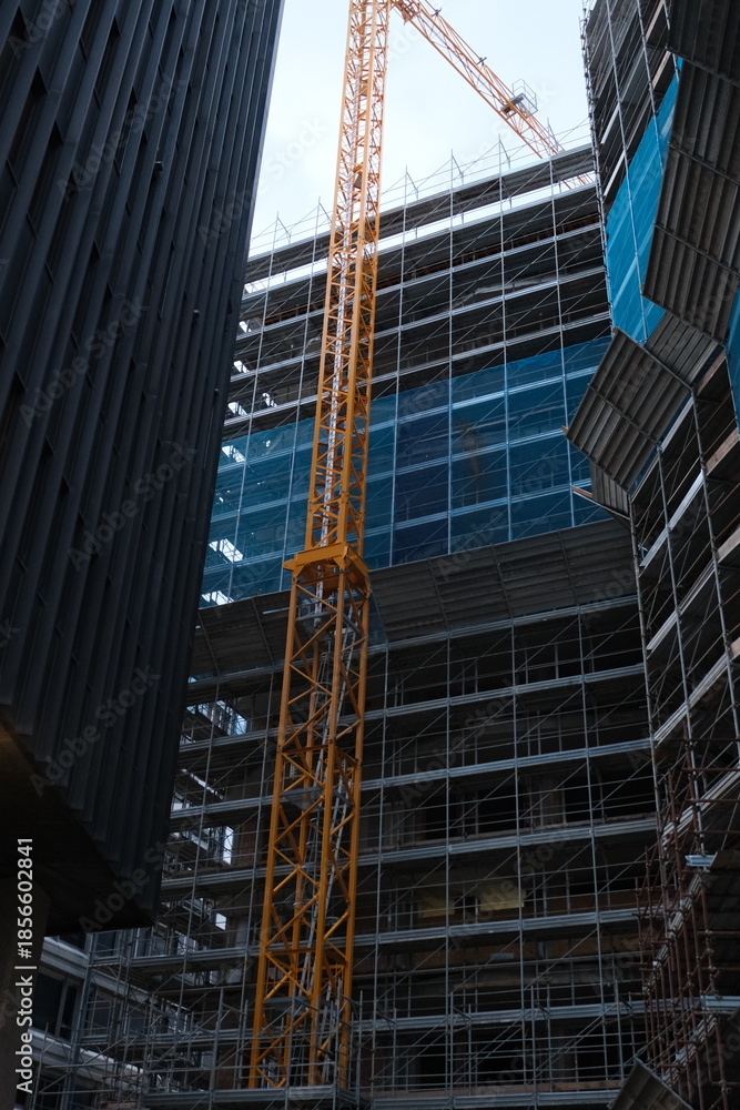 Fototapeta premium Urban construction site with scaffolding and yellow tower crane between modern buildings