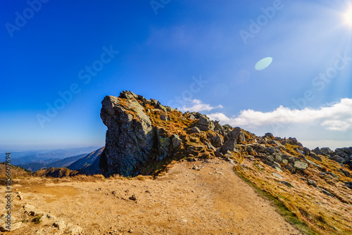A wide panoramic view of a rocky hiking trail winding through the slopes of the Low Tatras National Park during a sunny autumn day. Beautiful yellow grass and blue sky