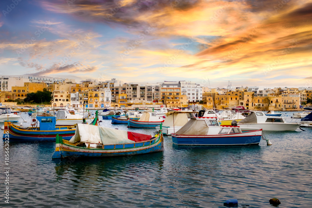 Obraz premium View of Birzebbuga's small harbor with numerous moored boats at sunset ; rows of houses appear in the background.