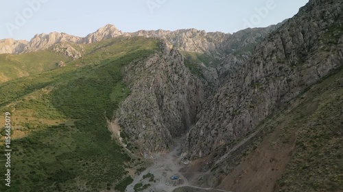 Aerial view of a narrow canyon carved between steep cliffs in Uzbekistan during early morning. Soft sun glows on green slopes and stone wall, the road and dry stream guide the eye through depth