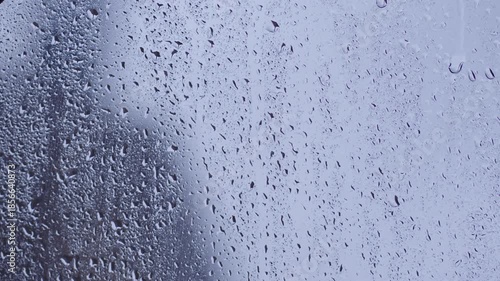 Closeup view of raindrops on transparent glass window during rainfall creating delicate patterns and reflections in blurred background