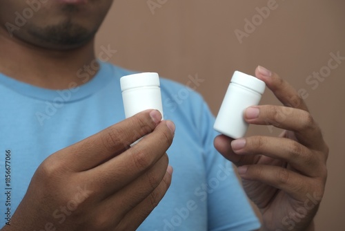 Close-up of Southeast Asian man holding two white pill bottles, representing medication choice, healthcare decision, and pharmaceutical concept for medical and advertising use.