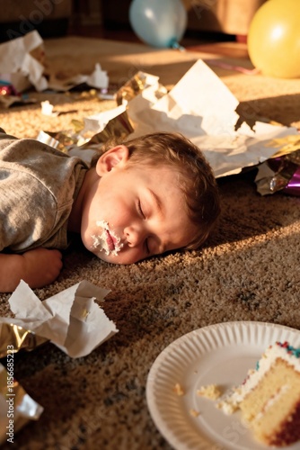 A toddler sleeps peacefully on a rug amid birthday chaos: torn wrapping paper, a balloon, and a half-eaten cake plate, a smear of frosting on their chin.
