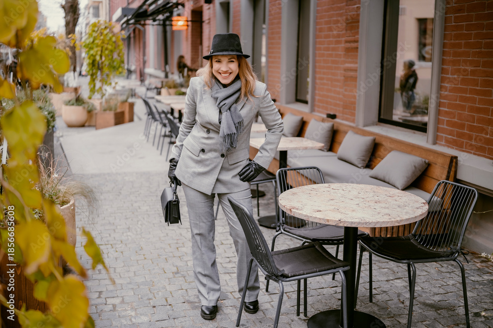 Obraz premium Smiling businesswoman in grey suit and black hat walking in cozy city street in late autumn, holding small handbag in gloved hand. Urban walk, fashion, lifestyle concept.