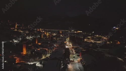 Night aerial view forwards of illuminated Mestia mountain ski resort village in winter with holiday decorations, snowy rooftops, and festive street ambiance