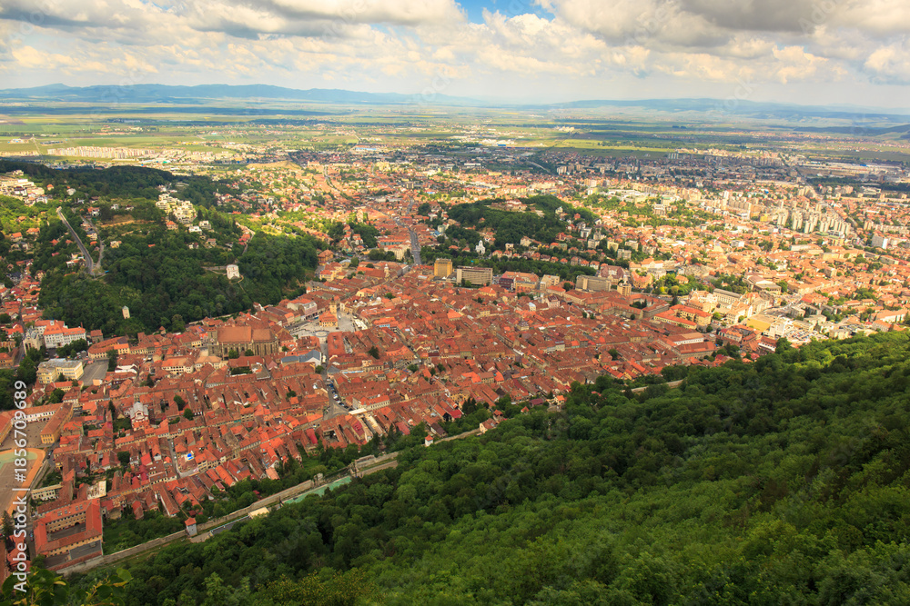 Fototapeta premium View from hill showing old and new Brasov with red rooftops, cityscape merging with green hills on a sunny day.