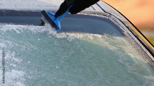 Individual using a blue ice scraper to remove frost from a car windshield, showcasing the action of clearing ice, with snow accumulation visible on the glass surface