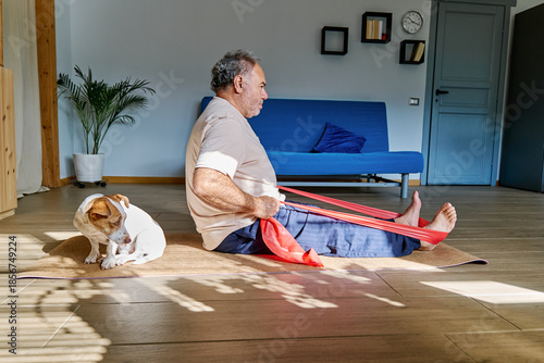 Mature man doing fitness training with resistance band on fitness mat with his Jack Russell Terrier dog at home. Healthy lifestyle for wellness.