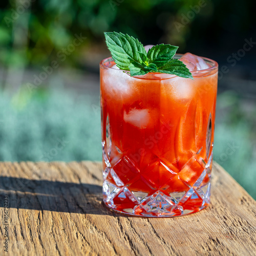 Bloody mary cocktail with ice cubes and green mint leaves on a nature background. Red tomato juice in glass on a wooden table on a sunny day, closeup