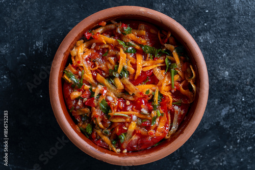 Delicious pickled tomatoes with carrots, garlic and parsley in a clay bowl on a dark background, closeup, top view