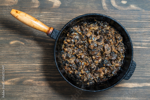 Cast iron skillet with fried wild mushrooms with onions and sour cream on wooden background, top view, closeup