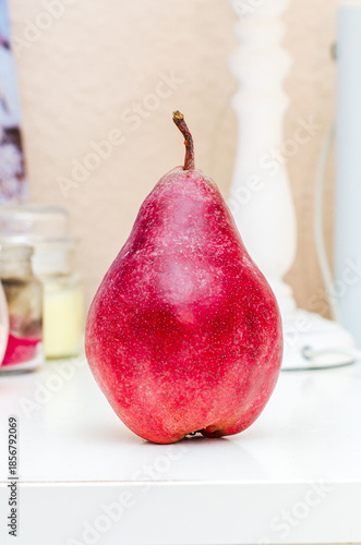 A single, vibrant red pear with a delicate stem sits on a clean white surface, its skin speckled with subtle variations in color and texture