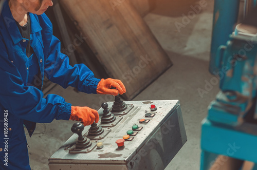 Bild auf Leinwand Industrial worker in blue uniform, orange gloves operating control panel with joysticks and buttons