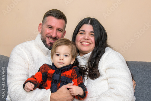 Smiling family portrait of two adults holding a toddler while seated together indoors.