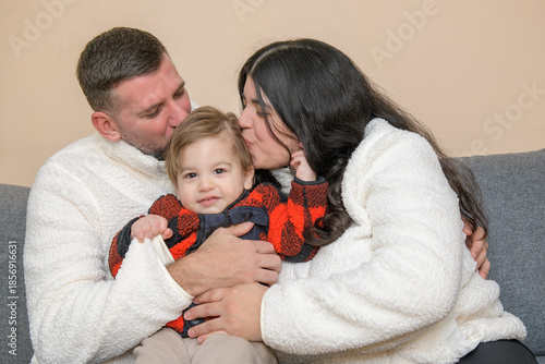 Parents kissing their toddler while sitting together on a couch indoors.