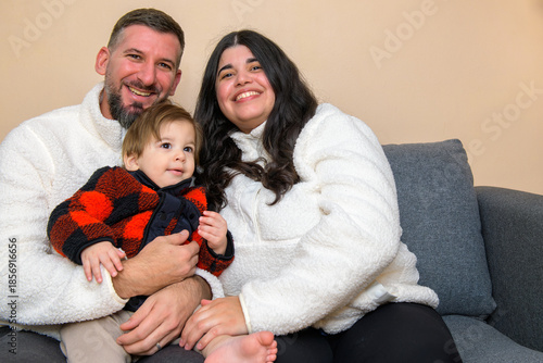 Smiling family sitting together on a couch, holding a toddler in a cozy indoor setting.