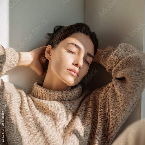 Woman relaxing in soft light against a wall with hands behind her head in a cozy indoor space during the day