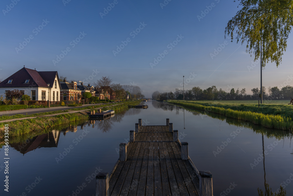 Obraz premium morning view from a wooden pier over the bedum canal. dutch landscape with mist, traditional houses and perfect reflection on the water