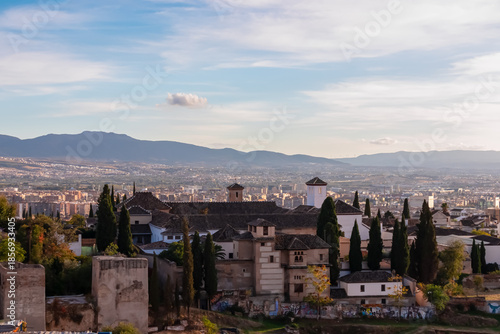 High angle view captures the historic buildings and cypress trees of the Alhambra complex overlooking the sprawling city of Granada and distant mountains under a soft evening sky in Andalusia, Spain.