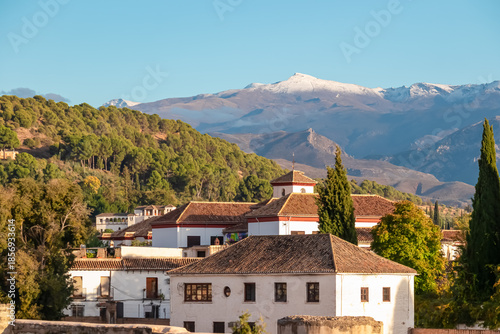 White traditional houses with tiled roofs stand against the snowy Sierra Nevada mountains in Granada, Spain, highlighting the contrast between the sunny city architecture and alpine peaks.