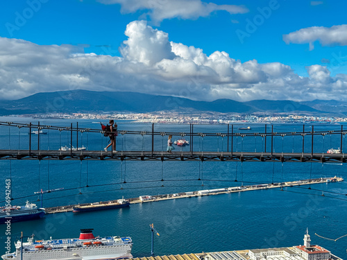 Tourists traverse the long span of the Windsor Suspension Bridge, silhouetted against a backdrop of the deep blue Bay of Gibraltar, large cargo ships, and the distant mountains of the Spanish coast.