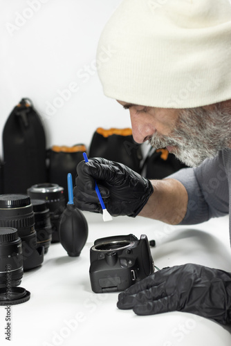 A bearded technician wearing gloves cleans a camera sensor using a specialized spatula, working carefully at a table surrounded by lenses and professional photography equipment.