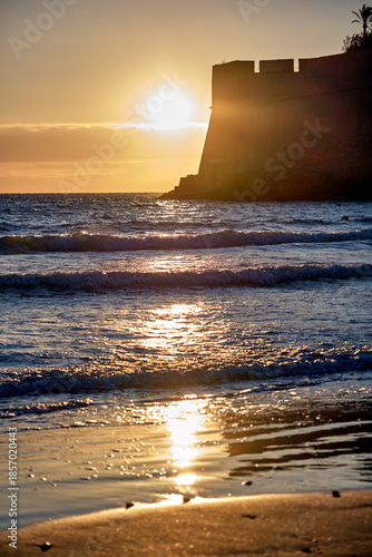 Sunset over Peñiscola beach with castle silhouette and waves reflecting light on water surface