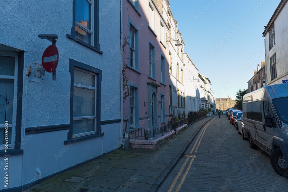 Fototapeta premium Narrow street lined with pastel-colored buildings, bright sunlight, no entry sign, parked vans, creating a quaint, peaceful, and slightly compressed perspective in Caernarfon - Gwynedd - Wales - UK