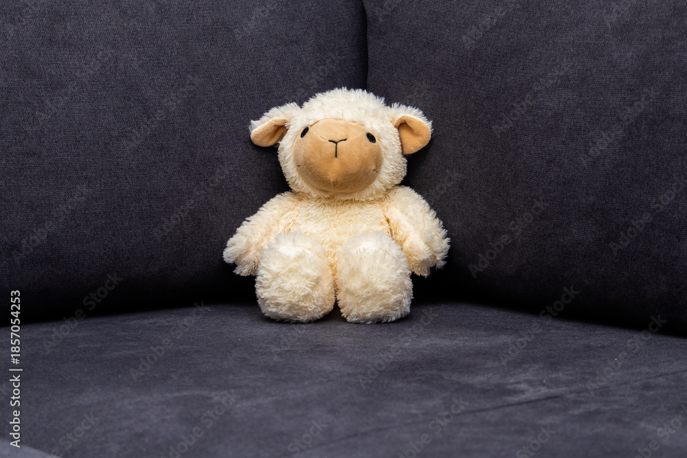 Fototapeta premium Cute plush lamb toy sitting on a dark sofa. Minimal cozy interior scene, childhood, comfort and nursery decor concept with copy space.