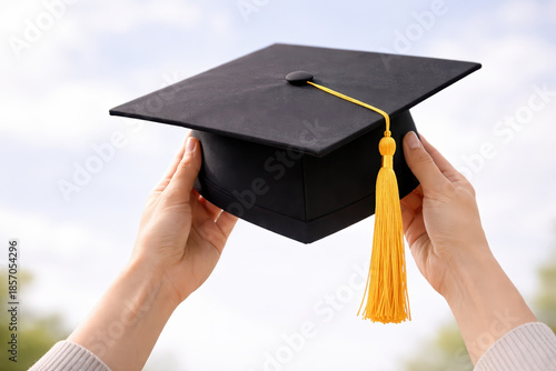 Graduation cap with yellow tassel held by hands against blurred outdoor background, symbolizing academic achievement and celebration of success