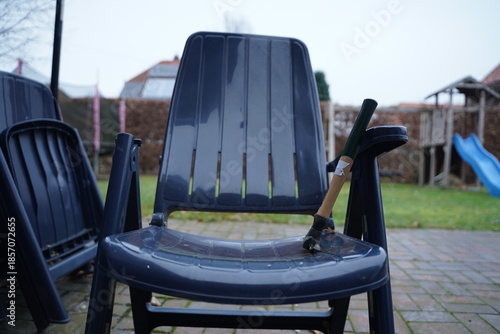 Dark plastic garden chair on a patio with a broken armrest and repair tape; other stacked chairs in the background. Outdoor furniture damage is shown.