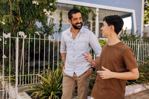 Father and teenage son smile while talking during a walk in a small residential neighborhood, showing connection, communication, emotional balance, and a healthy family relationship