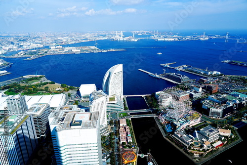 横浜みなとみらいの全景を高いビルから見下ろし青い海と近代的な建物や大きな観覧車が広がる絶景を捉えています。