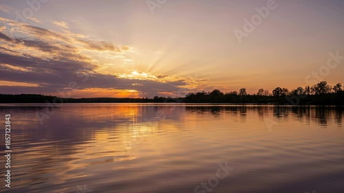 Serene Sunset Over Calm Lake with Dramatic Sun Rays Breaking Through Clouds and Gentle Ripples Reflecting Golden Hour Light and Silhouette