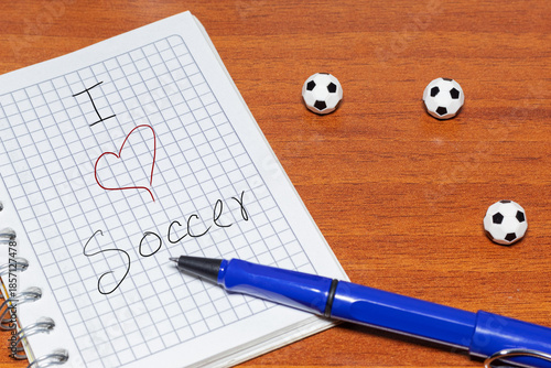 Notebook with the message “I love soccer” and a blue pen on a desk with miniature soccer balls. Concept of passion and sporting well-being