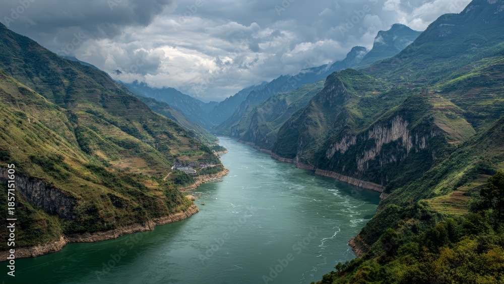 Obraz premium A wide deep river flows between steep green terraced mountains under a cloudy sky