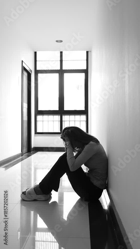 A lonely young woman sits on the floor in a narrow hallway, clutching her head in despair, expressing sadness, stress, and emotional conflict in the quiet, simple interior of the building. The image i