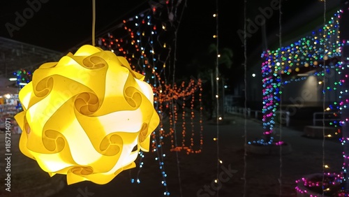 Illuminated Sphere in Festive Night: A radiant yellow sphere illuminates the night, surrounded by strings of colorful lights creating a festive atmosphere, against a backdrop of buildings. 