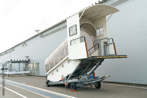 Portable aircraft boarding stairs with a protective canopy parked outside an airport hangar. Mobile ramp for airplane embarkation and disembarkation at a professional aviation facility.